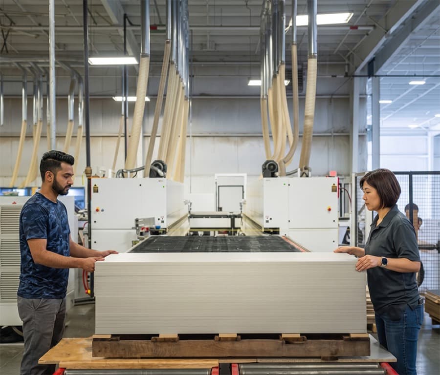 Two workers guiding a stack of panel stock onto a pallet beside a CNC line in a store-fixtures plant